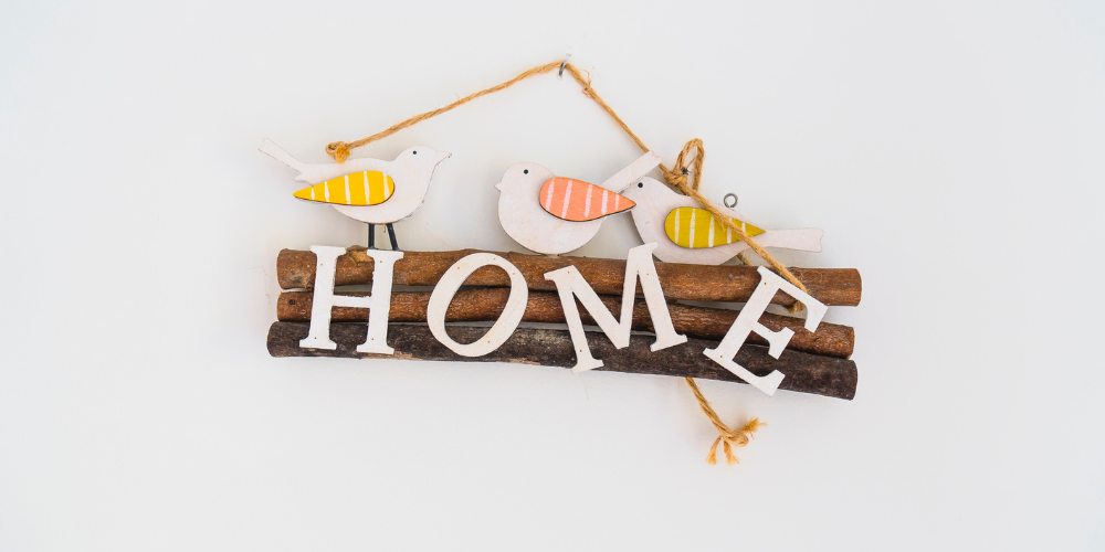 Wooden sign against a white wall. The sign says home with wooden birds on top.