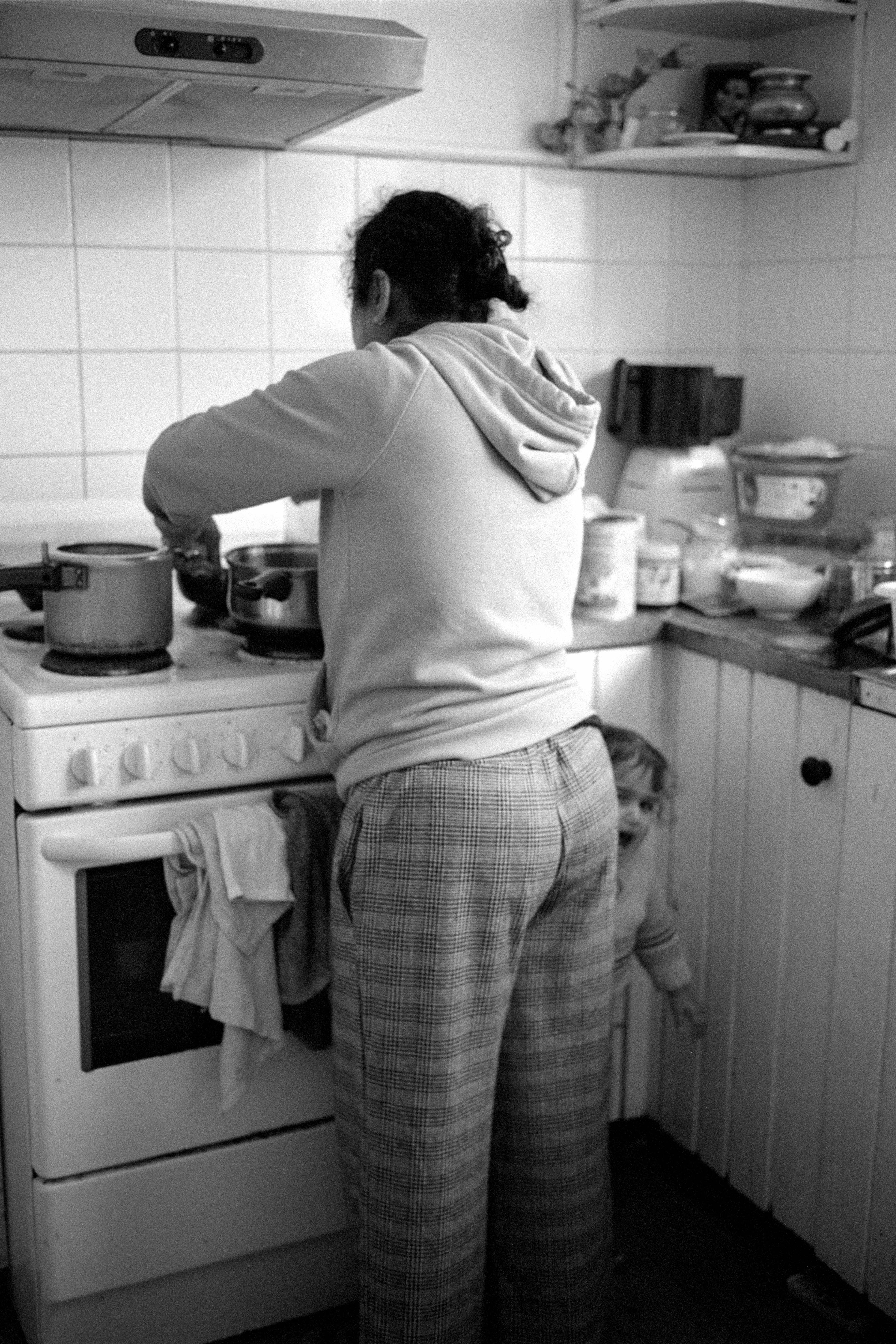 Black and white photograph of a person cooking over a stove in the kitchen, with a small child next to their leg