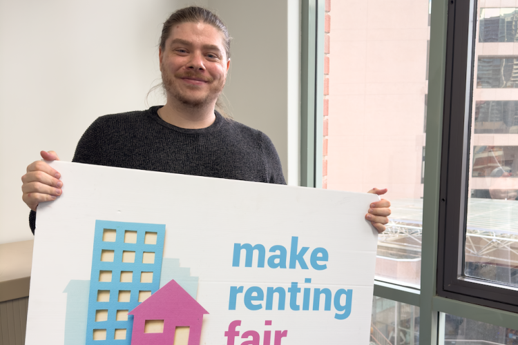 Dylan holding a sign saying make renting fair in front of a window.